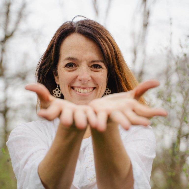 Gesangsunterricht Online Frau mit braunen Haaren, die lächelt und beide Hände mit offenen Handflächen ausstreckt.