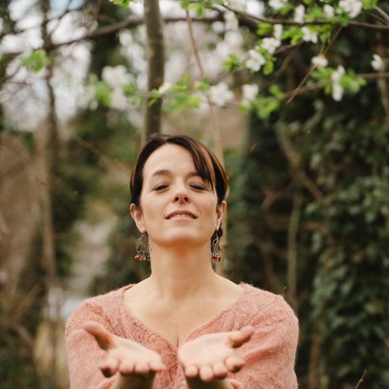 Monica Sardi Gesangslehrerin Frau mit offenen Händen steht in der Natur, umgeben von blühenden Bäumen.